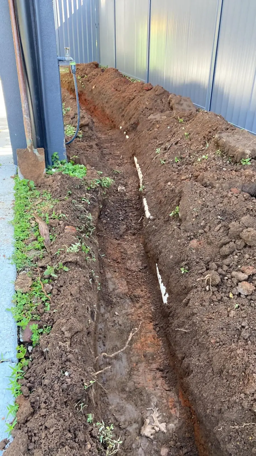 Freshly dug drainage trench along a side path between house and fence